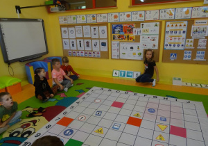 Czworo dzieci siedzi po lewej stronie maty. Dziewczynka układa przed matą kod z tabliczek ze strzałkami kierunkowymi według którego będzie programowany Bee bot.